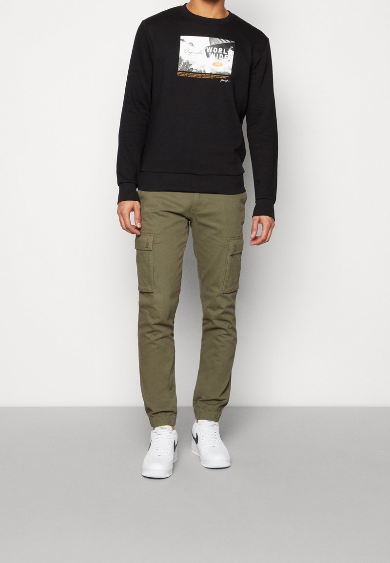 Felpa nera con collo girocollo e stampa grafica, abbinata a pantaloni cargo verde oliva con tasche laterali e sneakers bianche con dettagli neri.