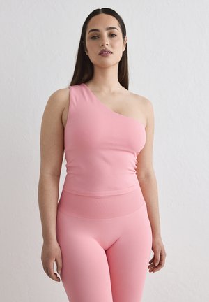 Femme aux cheveux raides et foncés portant un haut rose à une épaule et des leggings taille haute assortis, debout devant un fond blanc texturé.