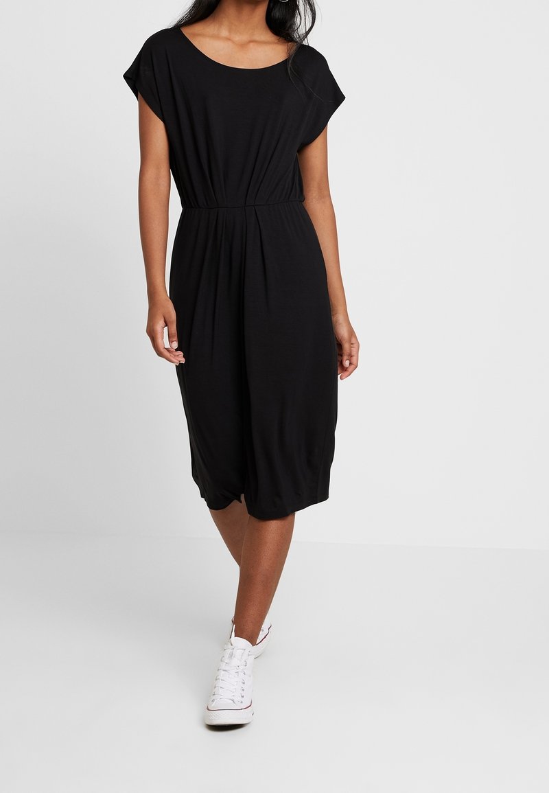 Femme portant une robe noire longueur genou avec une taille cintrée et des manches courtes, associée à des baskets blanches montantes, marchant vers l'avant.