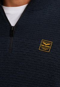 Marineblauwe trui met kwart-ritssluiting en een gestructureerd breipatroon, voorzien van een zwarte rits. Heeft een geel PME Legend merkembleem op de borst.