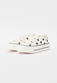 Białe płócienne trampki z czarnymi groszkami i czerwonym napisem, zaprojektowane z grubą białą podeszwą i czarnymi paskami akcentującymi.