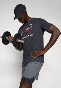 Svart Under Armour T-shirt med en röd logotyp, grå sportshorts och en svart keps; håller en svart hantel med texturerat grepp.