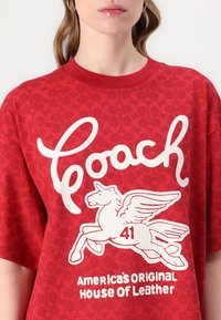 T-shirt vermelho de manga curta com um padrão texturizado, apresentando gráficos brancos de um cavalo e o texto "Coach 41 America's Original House of Leather."