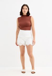 Top marrón sin mangas, pantalones cortos blancos con dobladillo ondulado y patrón recortado, combinados con sandalias deslizantes blancas. Textura suave y diseño ajustado.