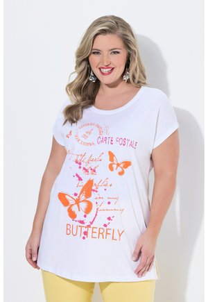 Hymyilevä nainen, jolla on aaltoilevat vaaleat hiukset, valkoinen T-paita, jossa oranssi perhoskuvio ja keltaiset housut yksiväristä taustaa vasten.