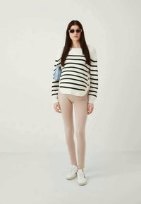 Sweater de malha listrado em preto e branco com botões, emparelhado com calças justas cor-de-rosa claro e ténis brancos. Modelo a segurar uma mala azul.
