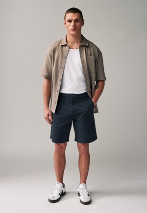 Jeune homme aux cheveux courts portant une chemise beige à manches courtes sur un débardeur blanc, un short bleu marine et des baskets blanches, se tient devant un fond uni.