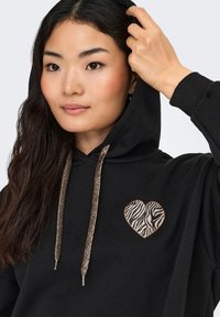 Sudadera con capucha negra que presenta un parche en forma de corazón con patrón de cebra en el pecho, con cordones metálicos dorados que acentúan el diseño.
