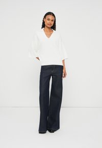 Suéter branco em V com mangas largas combinado com jeans de perna larga azul-escuro. O conjunto apresenta um corte limpo e moderno, com um tecido texturizado.