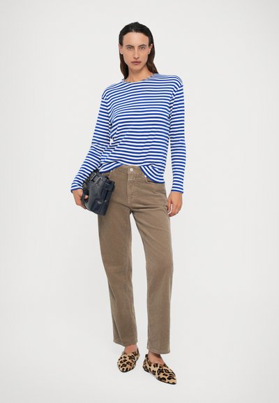 Vrouw in een blauw-wit gestreept longsleeve shirt, beige corduroy broek, lelijke muisjes in luipaardprint, met een zwarte handtas.