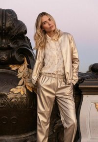 Veste bomber en satin doré associée à un pantalon en satin doré assorti. Le haut présente un chemisier texturé crème en dessous, créant un look superposé.