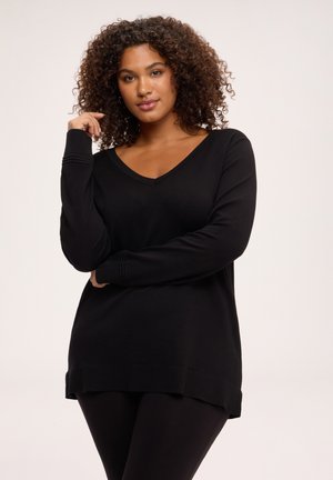 Femme aux cheveux bouclés portant un pull noir à col en V et manches longues ainsi qu'un pantalon noir, posant avec une main près du visage.