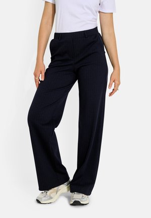 Mujer con pantalones anchos oscuros, camiseta blanca de manga corta y zapatillas plateadas, de pie con una rodilla ligeramente doblada.
