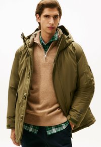 Veste matelassée vert olive avec capuche, dotée d'une fermeture éclair frontale et de plusieurs poches, portée par-dessus un pull beige et une chemise à carreaux verte.