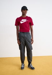 Camiseta Nike color marrón con logo blanco, gorra negra, pantalones cargo grises con puños elásticos y zapatillas negras con acentos blancos.