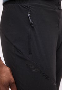 Κοντινό πλάνο ενός ατόμου που φοράει μαύρο παντελόνι με κάθετη τσέπη με φερμουάρ και διακριτική σήμανση "DYNAFIT" στο μηρό.