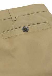 Gros plan sur la poche arrière d'un pantalon beige avec fermeture à bouton et passant de ceinture, montrant les détails des coutures et la texture du tissu.