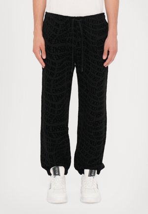 Black sweatpants feature a textured pattern of the word "FUTURE" in varying sizes. The waistband has a drawstring; worn with white sneakers.