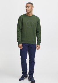 Zöld crewneck pulóver domború kör alakú logóval, tengerészkék nadrággal és fekete sportcipővel párosítva. Sima textúra, laza fazon.