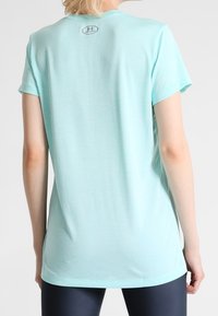 Person som bär en ljusblå kortärmad Under Armour-tröja, sedd bakifrån, tillsammans med mörka leggings mot en enkel bakgrund.