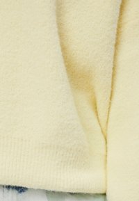 Tissu en laine douce jaune pâle avec une texture et des plis visibles, reposant sur une surface bleu et blanc faiblement motifée.