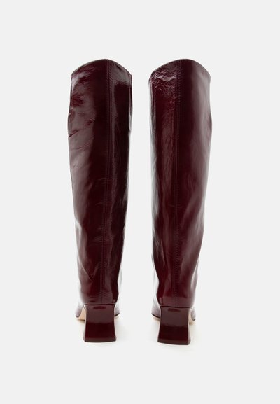 Ζευγάρι ψηλών, γυαλιστερών σκούρων μπορντό δερμάτινων μπότες με χοντρό τακούνι, από πίσω σε απλό λευκό φόντο.