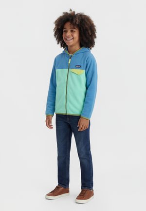 Niño sonriente con cabello rizado que lleva una sudadera con capucha y cremallera azul y verde, jeans oscuros y zapatillas marrones, de pie contra un fondo liso.