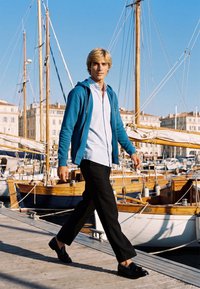 Jeune homme en sweat à capuche bleu et pantalon noir marchant sur un quai en bois avec des voiliers amarrés dans une marina ensoleillée.