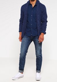 Marine gebreide cardigan over een donkerblauw overhemd met knopen, gecombineerd met medium-was jeans en witte sneakers. Kenmerkt zich door een geribbelde textuur en opgerolde mouwen.
