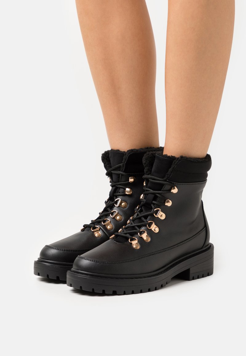 Bottines noires en cuir avec un col en fausse fourrure, œillets dorés et lacets noirs, dotées d'une semelle épaisse crantée pour une meilleure adhérence.