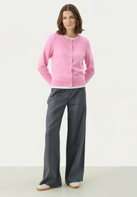 Cardigan rose clair avec un col rond et une fermeture par boutons, associé à un pantalon large gris et des baskets blanches. Texture lisse, design décontracté.