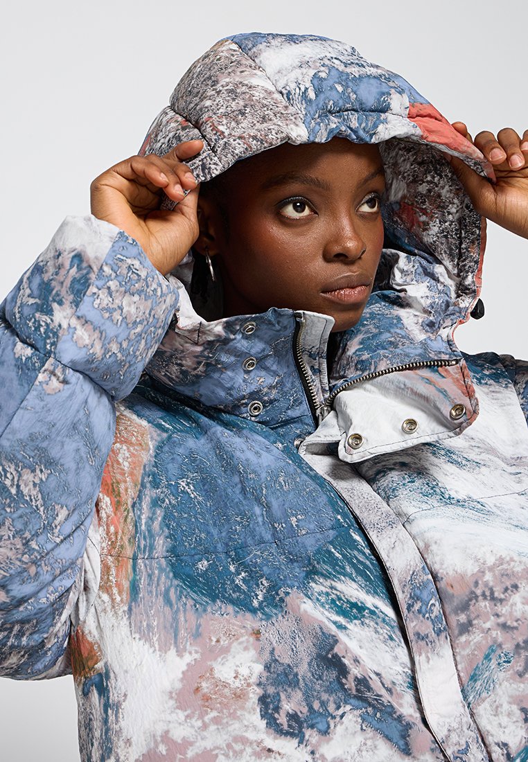 Veste matelassée avec un motif abstrait bleu, blanc et corail ; possède une capuche, des éléments en métal argenté et des fermetures à pression. Tissu chaud et texturé.