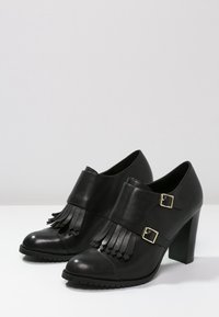 Zapatos de tacón alto de cuero negro con punta redondeada, detalle de flecos y dos hebillas doradas en una correa sobre la parte superior. Suela texturizada.
