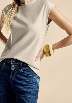 Kobieta w beżowej, bezrękawnikowej bluzce, niebieskich dżinsach i z dwoma dużymi, bursztynowymi bransoletkami na prawym nadgarstku na beżowym tle.