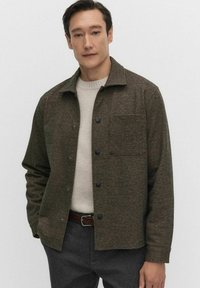 Bruine textuurjas met een knoopsluiting aan de voorkant, een enkele borstzak en een kraag, gedragen over een lichtcrème trui.