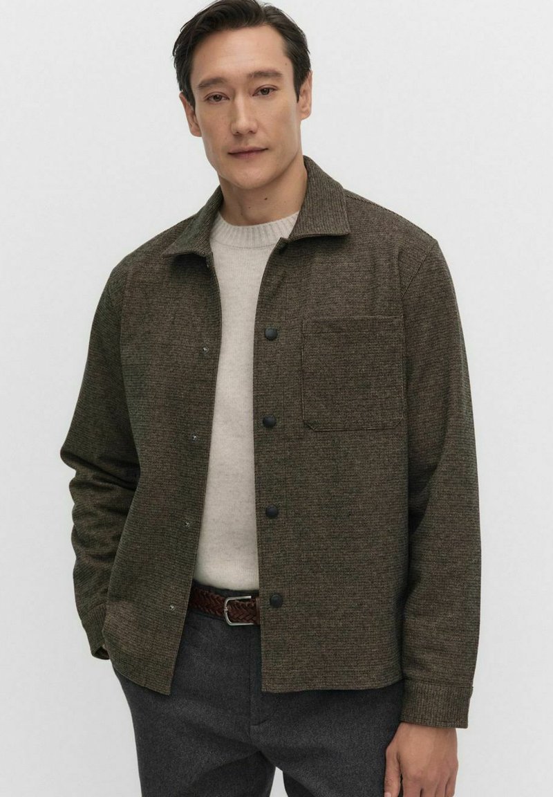 Bruine textuurjas met een knoopsluiting aan de voorkant, een enkele borstzak en een kraag, gedragen over een lichtcrème trui.