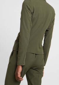 Veste ajustée couleur vert olive avec une texture lisse, manches longues et détails de coutures, assortie à un pantalon avec poches latérales.