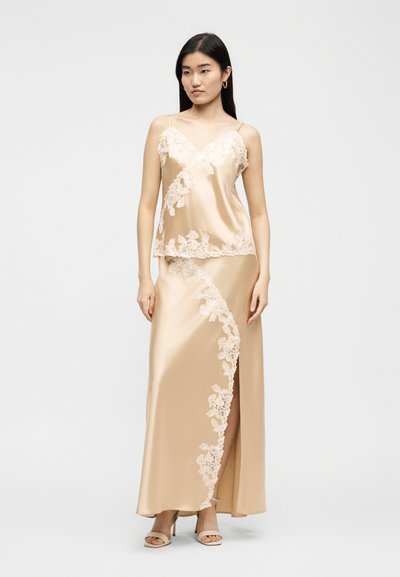 Γυναίκα με μπεζ σατέν καμιζόλ και μακρύ φόρεμα με λεπτομέρειες από φλοράλ δαντέλα και σκίσιμο μπροστά, στέκεται μπροστά σε απλό φόντο.