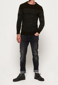 Tumma vihreä neulepusero, jossa tekstuuri kuvio, pyöreä kaula-aukko ja kangasmerkki. Yhdistetty käärityihin tummiin farkkuihin ja mustiin saappaisiin.