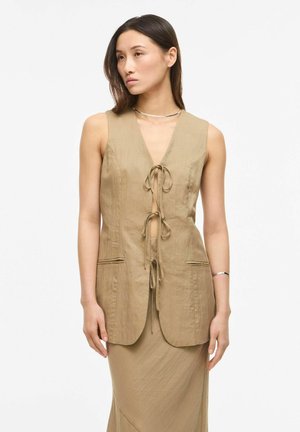 Donna che indossa un gilet beige senza maniche legato con tre fiocchi davanti sopra una gonna lunga abbinata, in piedi davanti a uno sfondo bianco.