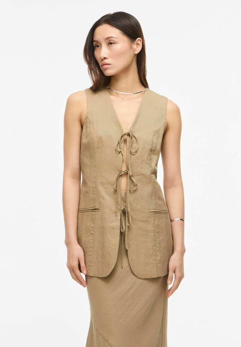 Femme portant un gilet beige sans manches noué avec trois nœuds à l'avant sur une jupe longue assortie, debout devant un fond blanc.