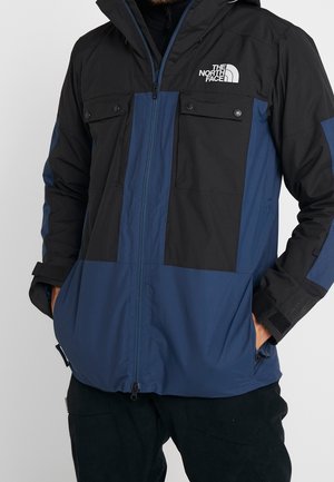 Czarna i ciemnoniebieska zimowa kurtka z kapturem, zamkiem z przodu, kieszeniami na klatce piersiowej i logo The North Face na piersi noszona przez osobę.