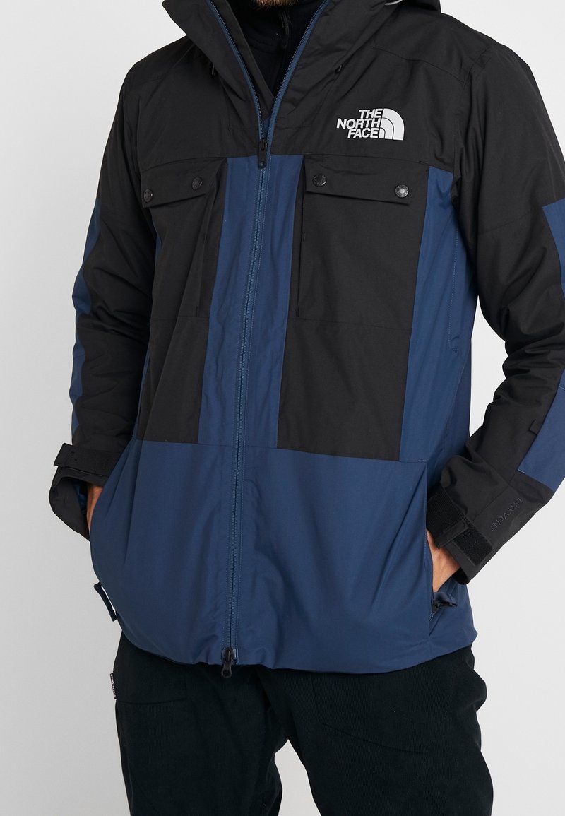 Veste d'hiver noire et bleu foncé à capuche avec fermeture éclair frontale, poches poitrine et logo The North Face sur la poitrine portée par une personne.
