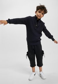 Zēns, ģērbies tumšā adītā džemperī un melnās biksēs ar daudzām kabatām, līdzsvaro ar izstieptām rokām, stāvot uz vienkārša gaiša fona.