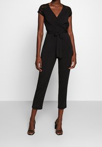 Combinaison noire avec des manches courtes en dentelle, un design enroulé et une taille à nouer. Présente des jambes fuselées et un tissu lisse. Portée avec des talons noirs.