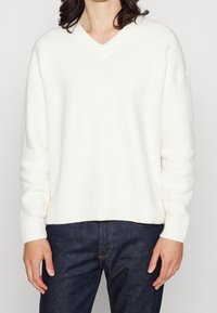 Bílý svetr s výstřihem do V z měkkého materiálu s žebrovanými lemy na rukávech a spodním okraji, doplněný tmavě modrými džínami. Jednoduchý a ležérní design.