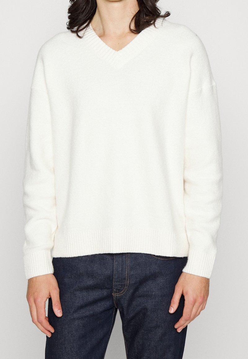Bílý svetr s výstřihem do V z měkkého materiálu s žebrovanými lemy na rukávech a spodním okraji, doplněný tmavě modrými džínami. Jednoduchý a ležérní design.