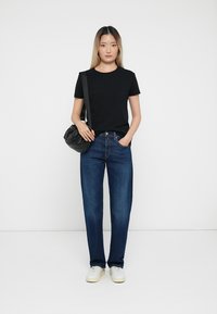 Zwarte katoenen t-shirt, blauwe denim jeans, witte sneakers en een kleine zwarte handtas. Model staat op een effen achtergrond.