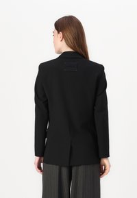 Černý blazer se strukturovanými rameny a dlouhými rukávy. Obsahuje centrální větrací otvor vzadu a obdélníkovou etiketu s červenými akcenty.