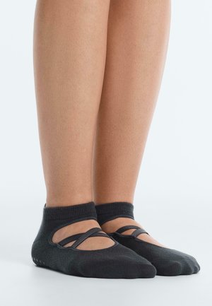 Lábak fekete, csúszásmentes balett zokniban, keresztpántokkal a felső részén, egyszínű világos háttér előtt.